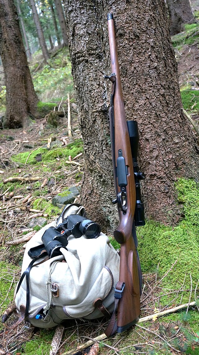 Blick auf einen Rucksack aus Leinen, ein darauf liegendes Fernrohr und eine Jagdwaffe, die an einem Baum im Wald lehnt. Untergrund: grünes Moos