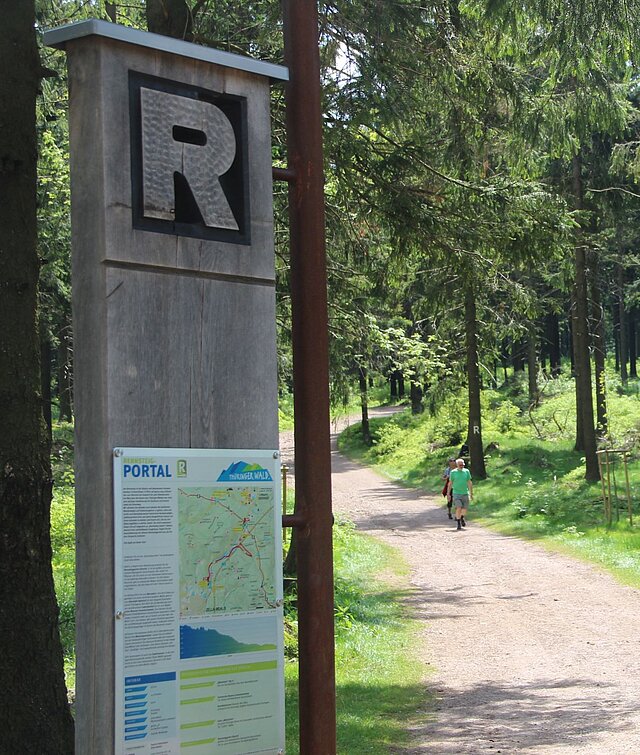 Ein Holzschild am Rennsteig mit dem typischen großen geschnitzten "R". Unter dem R sind Informationen zu den Wanderwegen und eine Karte angebracht.