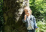 Kleines Mädchen mit lockigen Haaren und Zopf ateht im Wald an einem Baum und genießt ein Eis. Die Sonne scheint.
