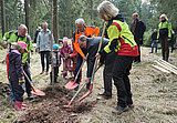 Gruppe von forstlich gekleideten Personen und Kindergartenkindern graben Löcher in den Waldboden, um junge Bäumchen zu pflanzen